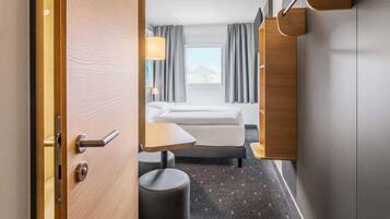 Ett hotellrum med en säng, ett litet bord, en stol, en rund pall och ett fönster med gardiner.