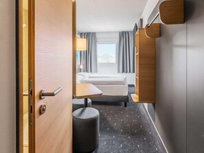 Une chambre d’hôtel avec un lit, une petite table, une chaise, un pouf rond et une fenêtre avec des rideaux.