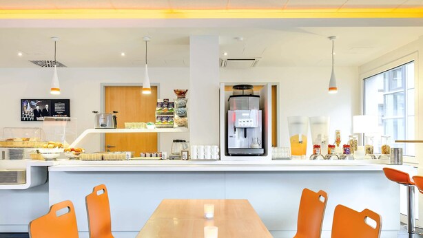 Un café moderno con una larga mesa de madera, sillas anaranjadas, una máquina de café y diversos alimentos en la barra.