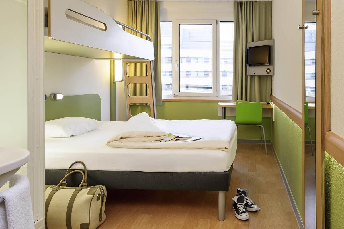 A hotel room with a bunk bed, a desk, a chair, a TV, and a window with curtains.