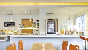 Uma área de café da manhã moderna com uma mesa de madeira comprida, cadeiras laranja, uma máquina de café e diversos alimentos.