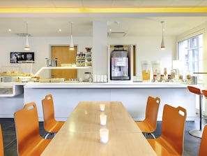 A modern breakfast area with a long wooden table, orange chairs, a coffee machine, and a variety of food items. - Ibis Budget Aachen City (Aachen)