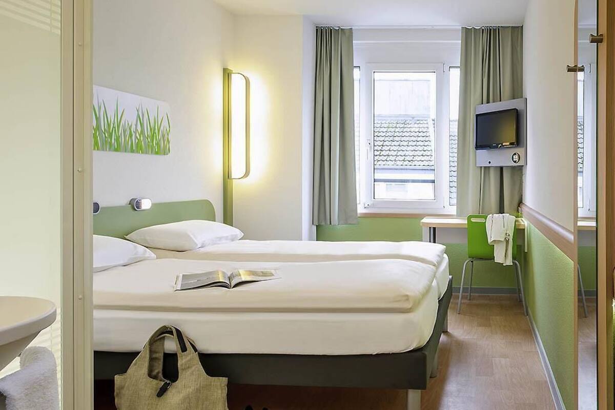 A hotel room with a bed, a bedside table, a TV, a window with curtains, and a view of buildings outside.
