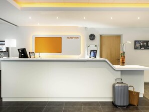 A reception desk with a digital clock, a first aid sign, and a framed picture on the wall. - Ibis Budget Aachen City (Aachen)
