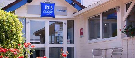 Un hotel con fachada azul y blanca, un letrero que dice 'ibis budget hotels', y un pequeño área exterior con sillas de metal.