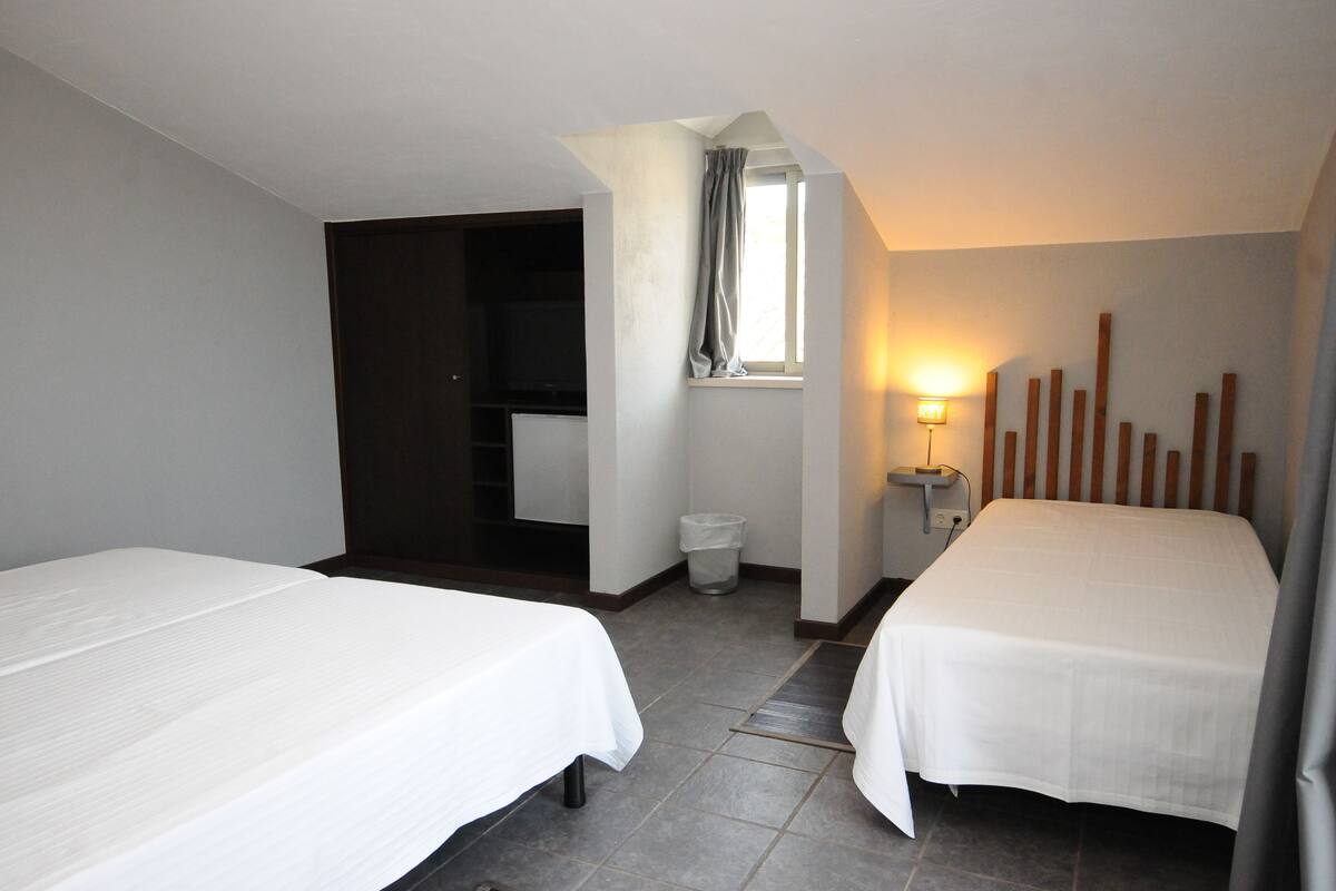A hotel room with two beds, a window with curtains, a small bedside table with a lamp, and a trash bin.