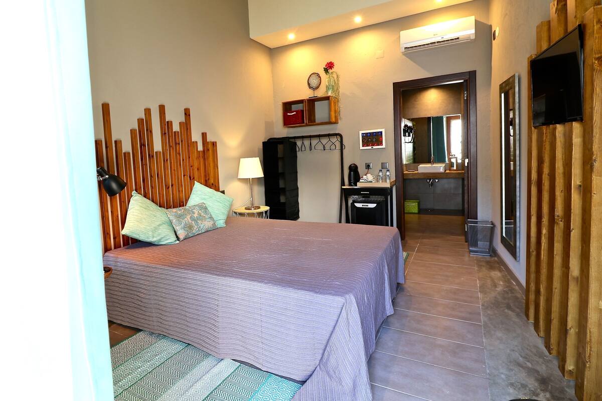 A bedroom with a bed, bedside table, lamp, and a wooden headboard. There is a bathroom visible through an open door.