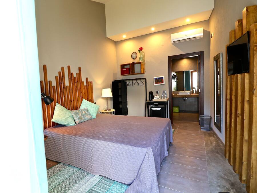 A bedroom with a bed, bedside table, lamp, and a wooden headboard. There is a bathroom visible through an open door.