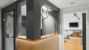 A reception desk with a B&B Hotels sign, a potted plant, and a modern interior with a flat-screen TV and seating area. - B&B HOTEL Flensburg-City (Flensburg)