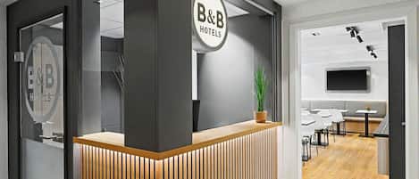 Una recepción con un letrero de B&B Hotels, una planta en maceta, y un interior moderno con televisor de pantalla plana y zona de estar.