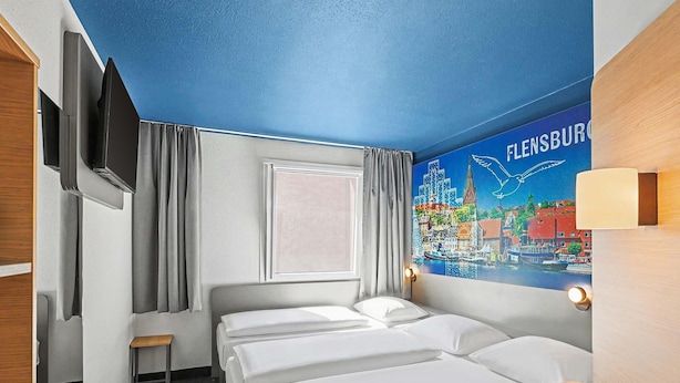 A hotel room with a bed, a TV, a window with curtains, a cityscape mural, and a wooden desk. - B&B HOTEL Flensburg-City (Flensburg)