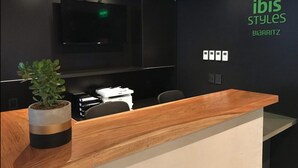 Un área de recepción con un mostrador de madera, una planta en maceta, un televisor de pantalla plana montado en la pared y un letrero que dice "ibis Styles BIARRITZ".