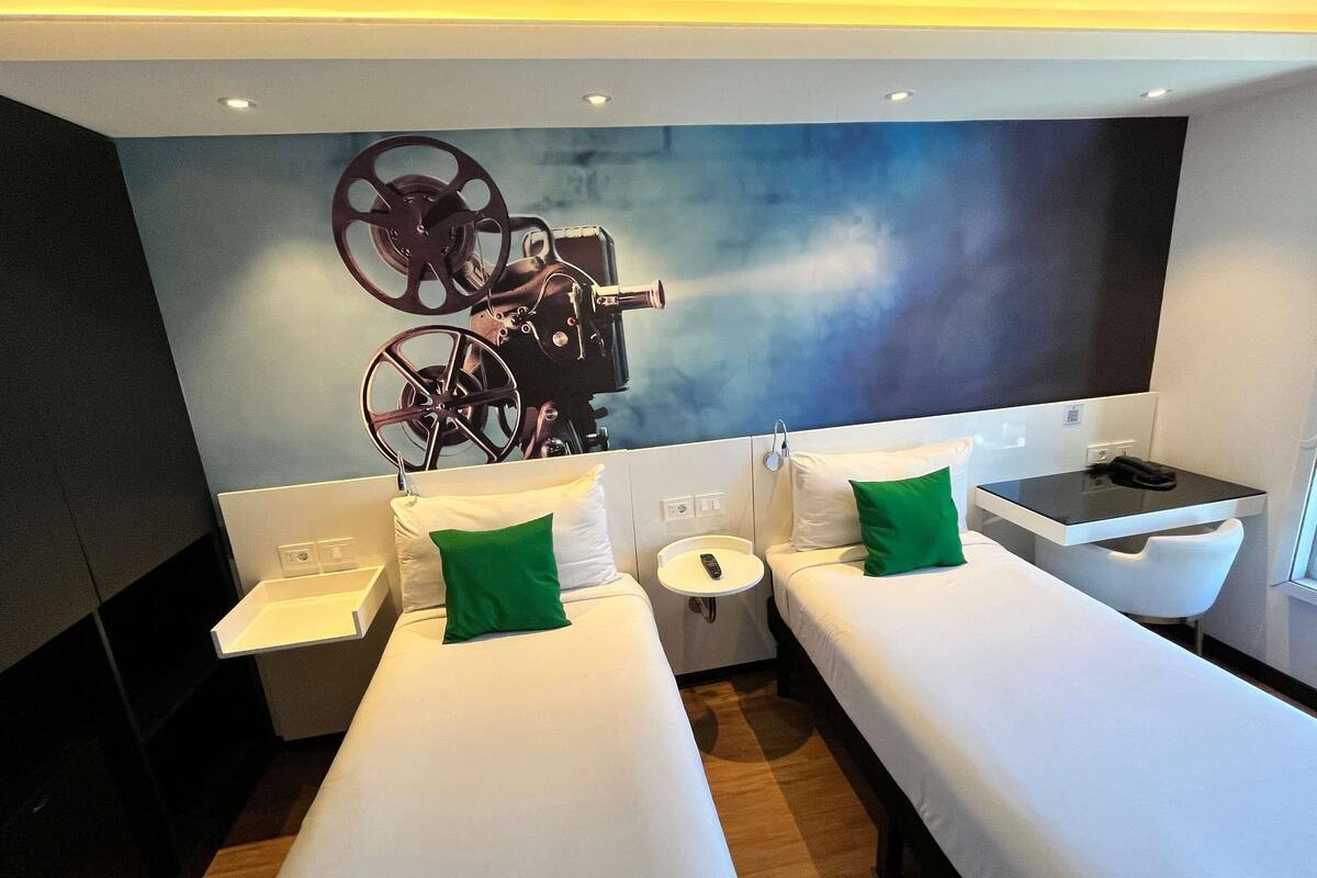 A hotel room with two single beds, a small table, a chair, and a large wall-mounted TV.