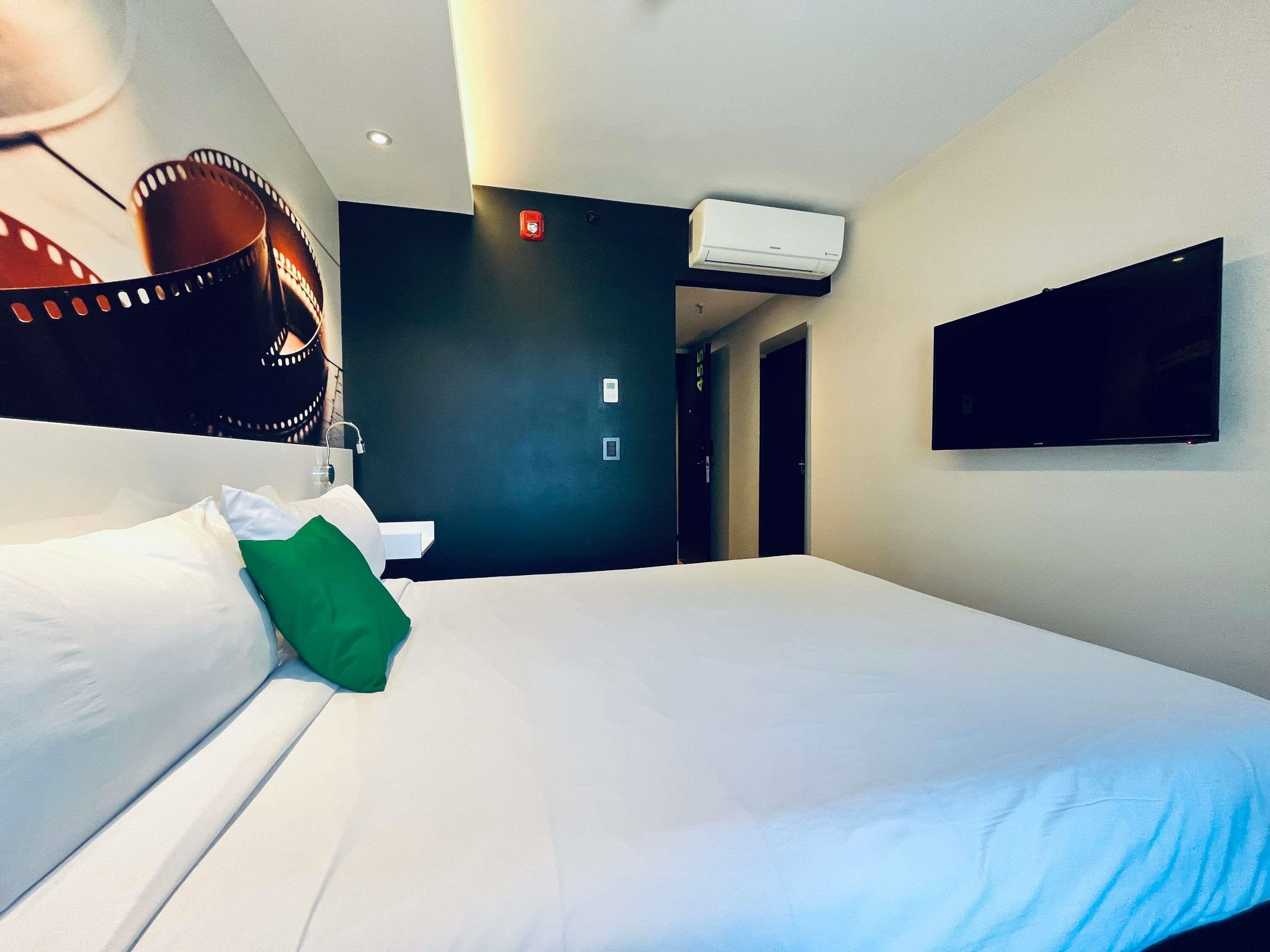 Habitación de hotel con una cama grande, un televisor de pantalla plana montado en la pared, un equipo de aire acondicionado sobre la cama y una puerta que da a otra habitación.