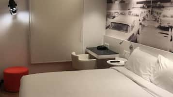 A modern hotel room with a large bed, a small desk, a red ottoman, and a wall-mounted air conditioning unit.