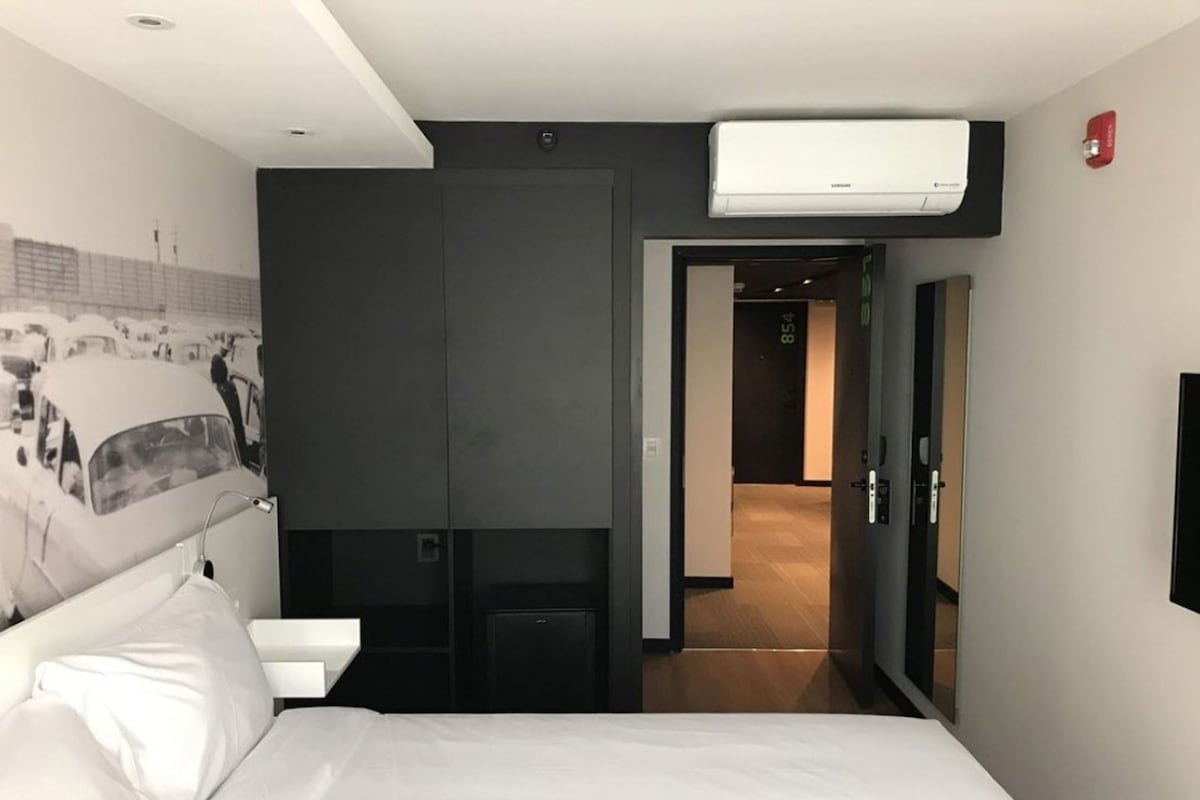 Una habitación de hotel moderna con una cama grande, aire acondicionado de pared, una puerta que da a un pasillo y un pequeño escritorio con una silla.
