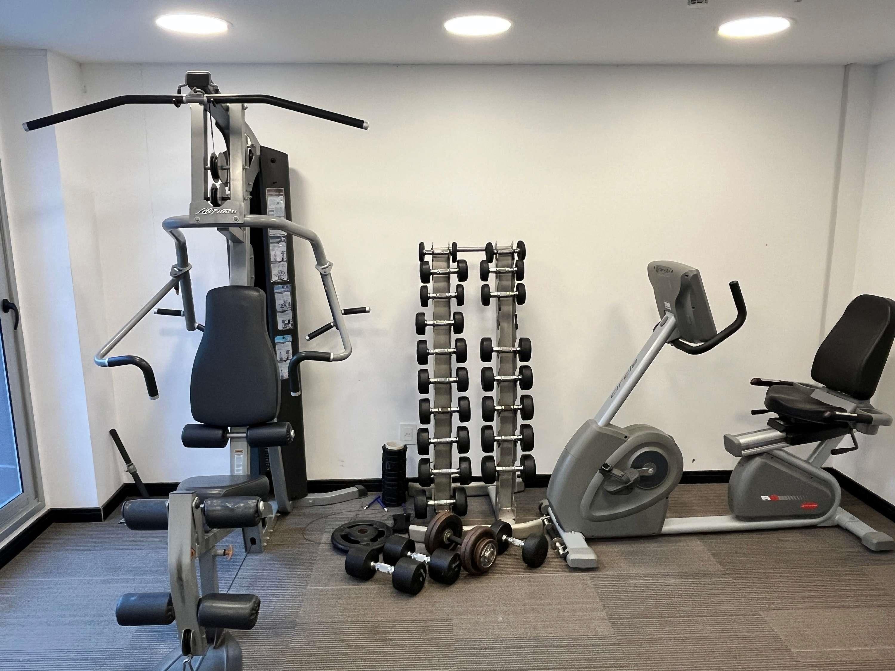 Un gimnasio con equipamiento de ejercicio, incluyendo un banco de pesas, mancuernas, una máquina de poleas y una elíptica.