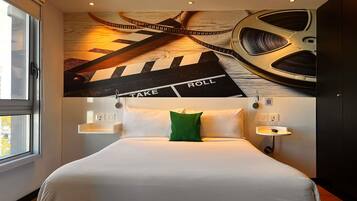 A hotel room with a bed, a green pillow, a wall-mounted clock, and a ceiling with a film strip design.