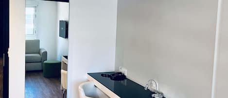 Una habitación moderna con escritorio, silla y un taburete pequeño. Hay una puerta que da a otra habitación con sofá y televisor.