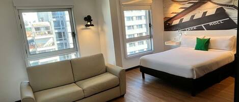A modern hotel room with a bed, a sofa, and two windows.