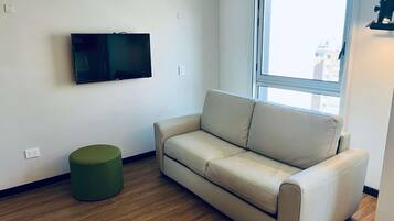 Una sala de estar con un sofá color beige, un otoman verde, un televisor de pantalla plana montado en la pared y una ventana que ofrece vistas de edificios exteriores.