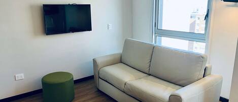 A living room with a beige sofa, a green ottoman, a flat-screen TV mounted on the wall, and a window providing a view of buildings outside.