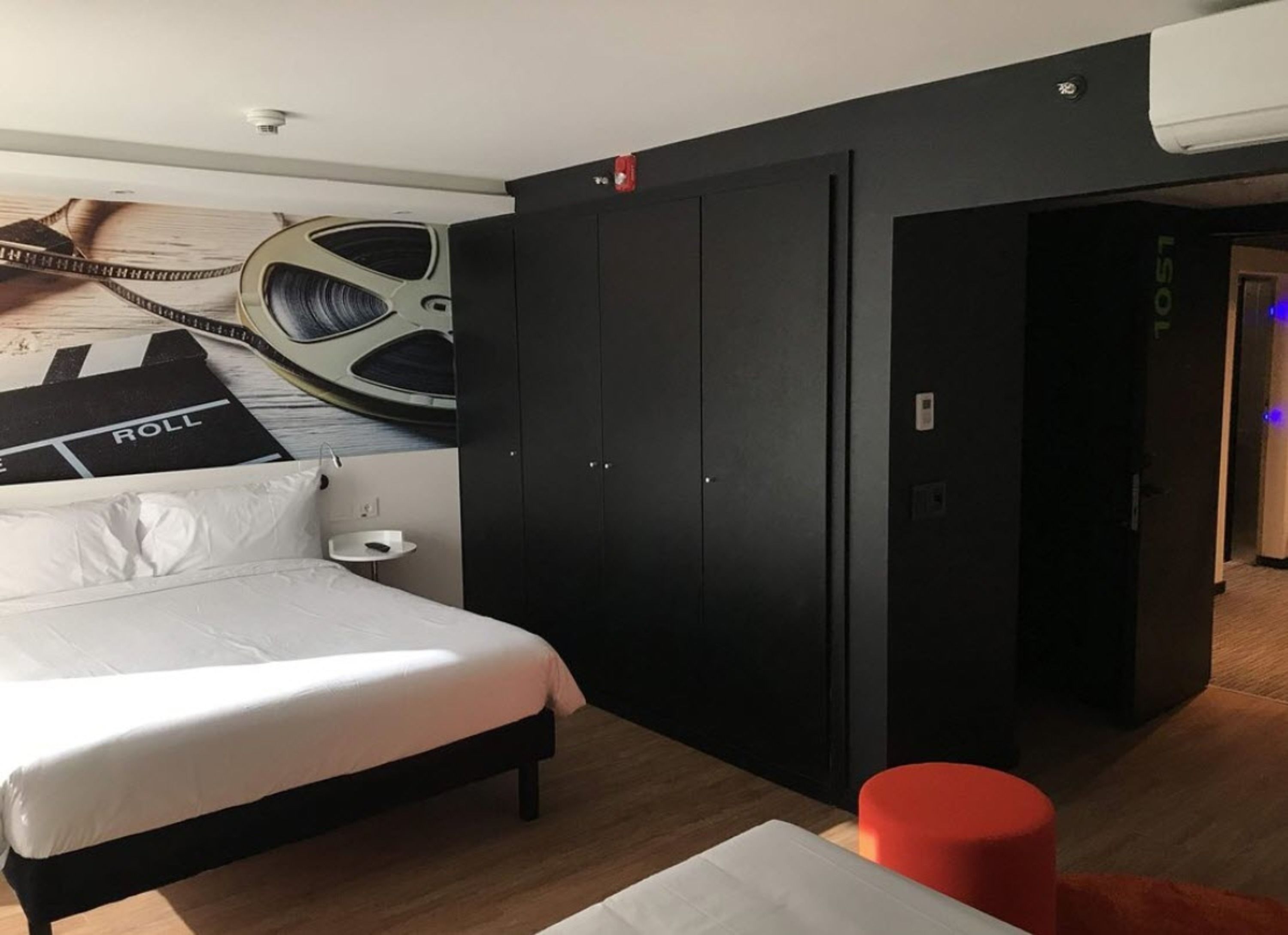 A modern hotel room with a bed, a wardrobe, a small bedside table, and a red ottoman.