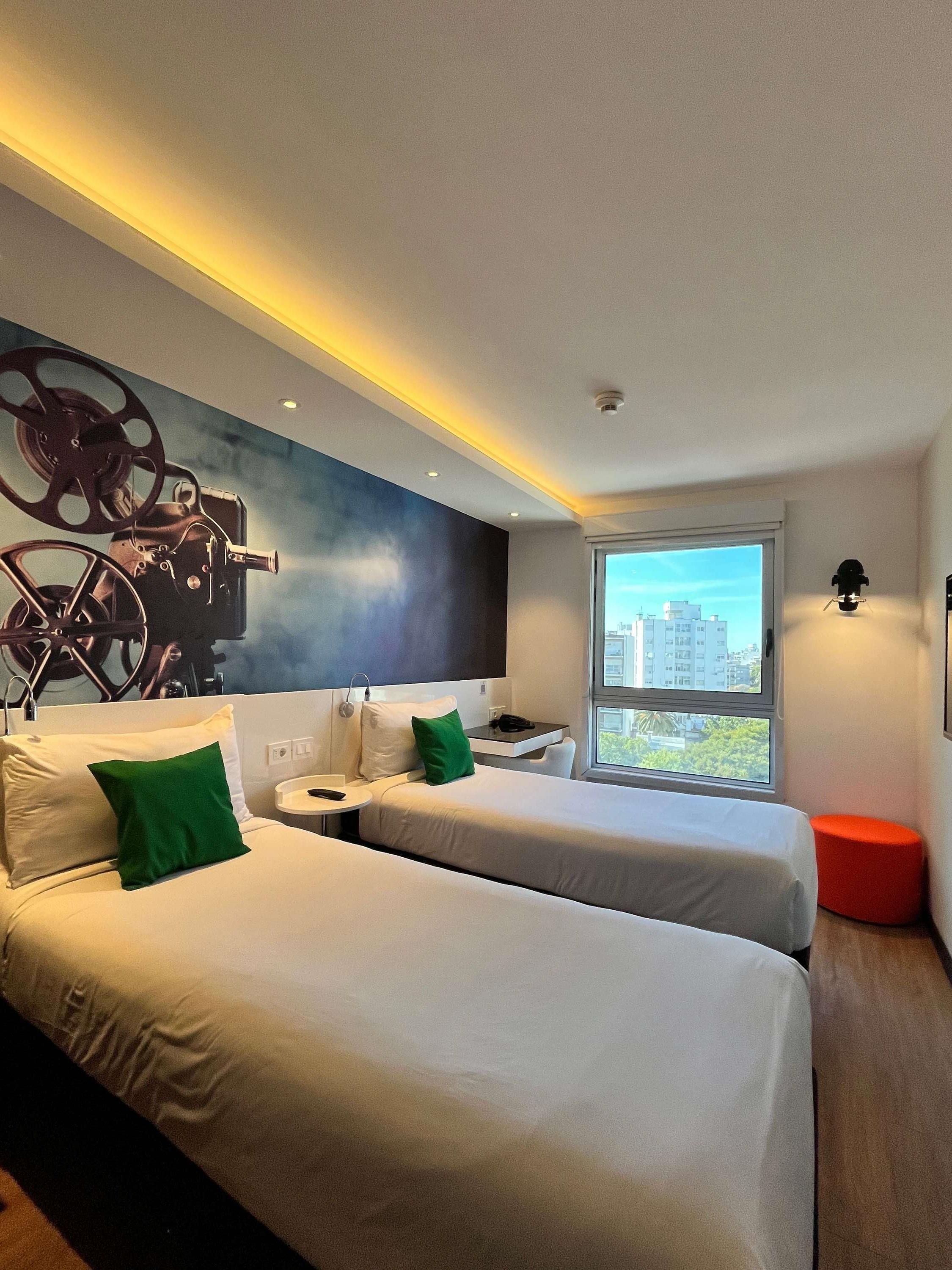 A modern hotel room with two beds, a small table, and a view of the city.