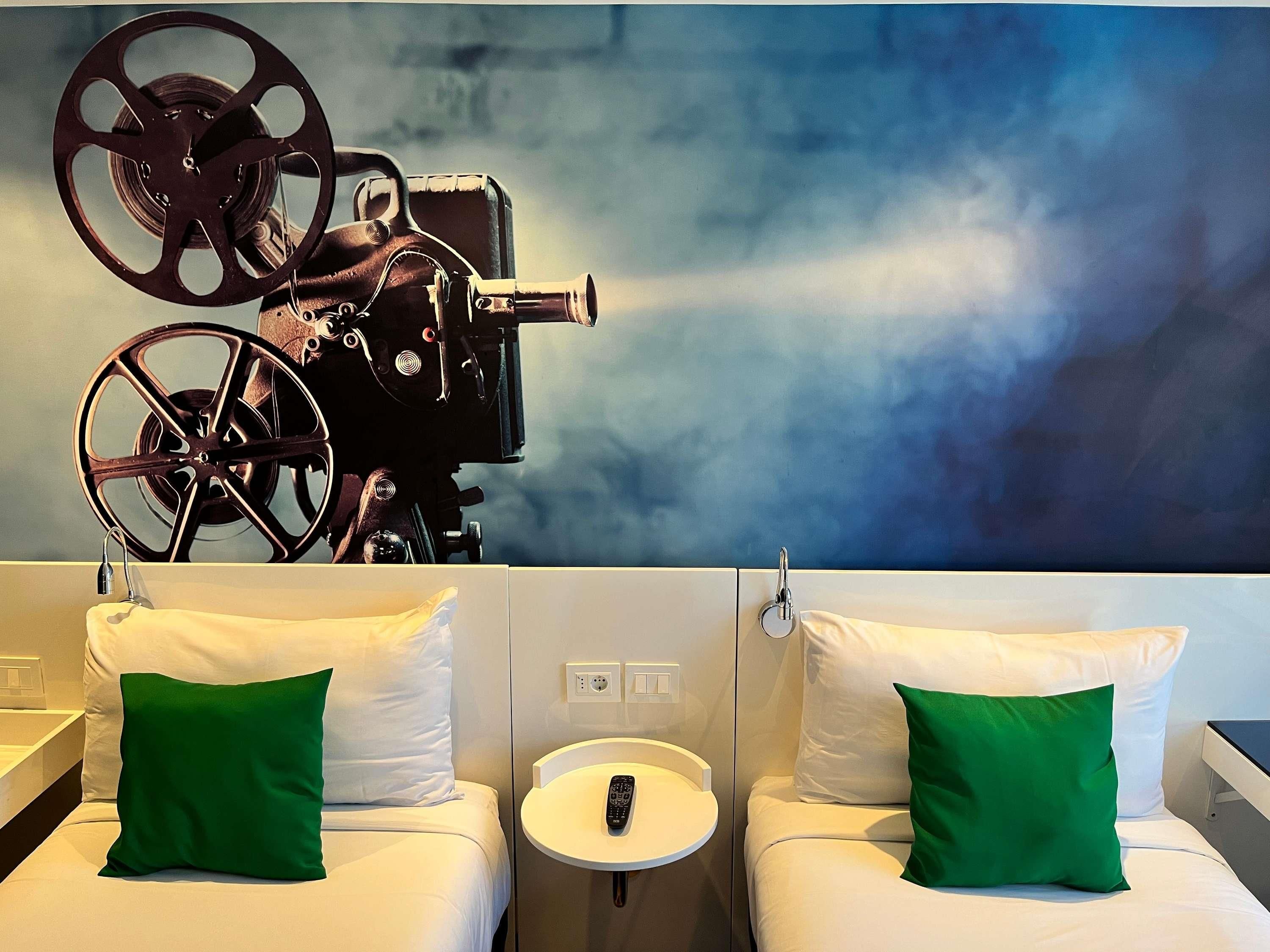 A hotel room with two beds, each with white bedding and green pillows. There is a round table with a remote control on it. A large movie projector is mounted on the wall above the beds.