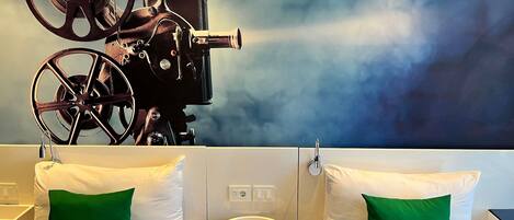 A hotel room with two beds, each with white bedding and green pillows. There is a round table with a remote control on it. A large movie projector is mounted on the wall above the beds.