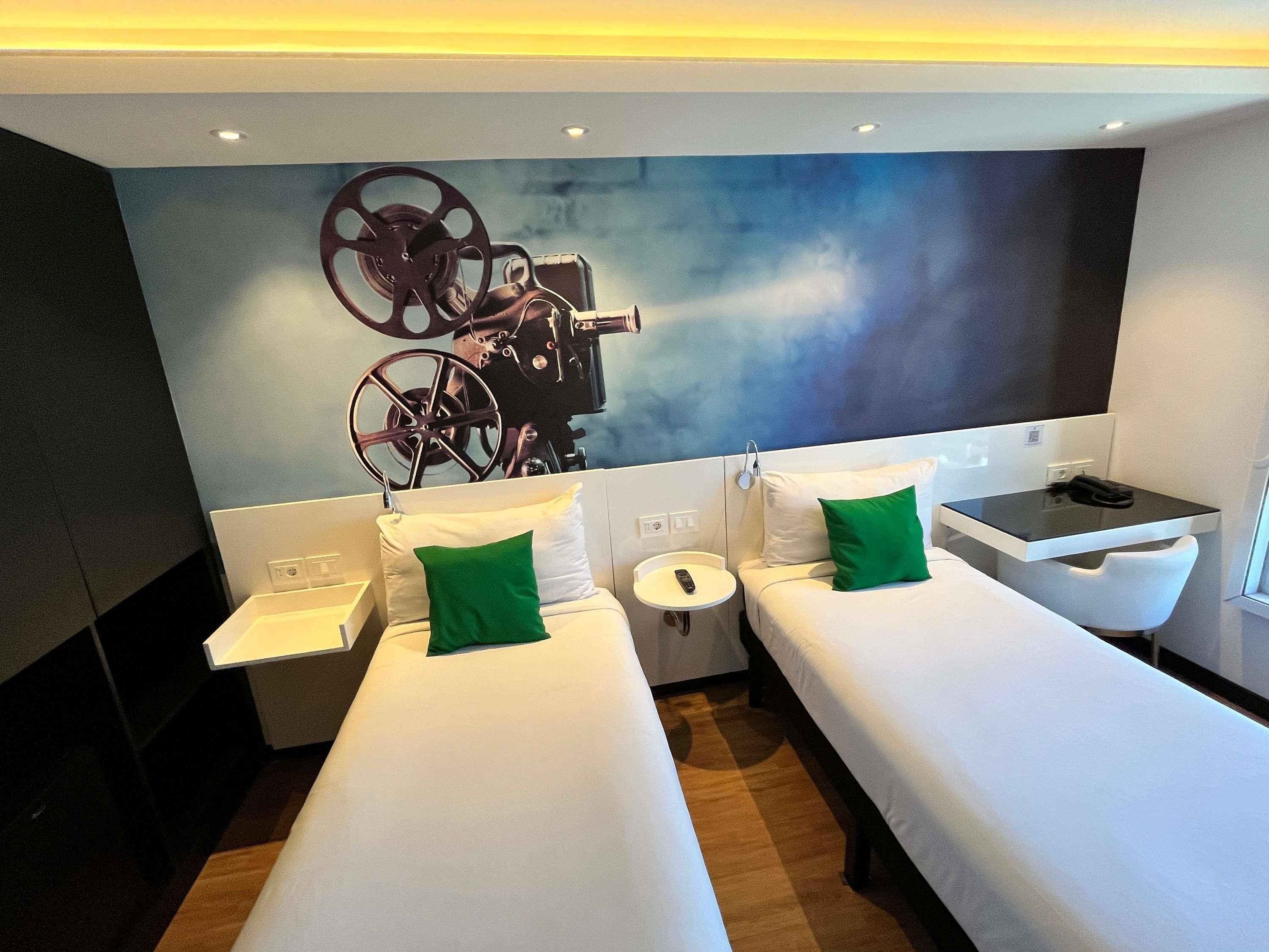 A hotel room with two single beds, a small table, a chair, and a large wall-mounted TV.