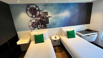 A hotel room with two single beds, a small table, a chair, and a large wall-mounted TV.