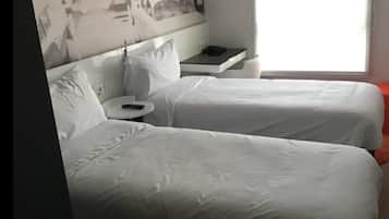 Habitación de hotel con dos camas, una mesita, una silla y ventana con vistas a un edificio.