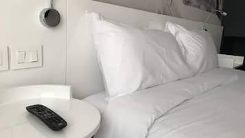Een hotelkamer met een bed, witte kussens, een ronde witte tafel, een afstandsbediening en een wandlamp.