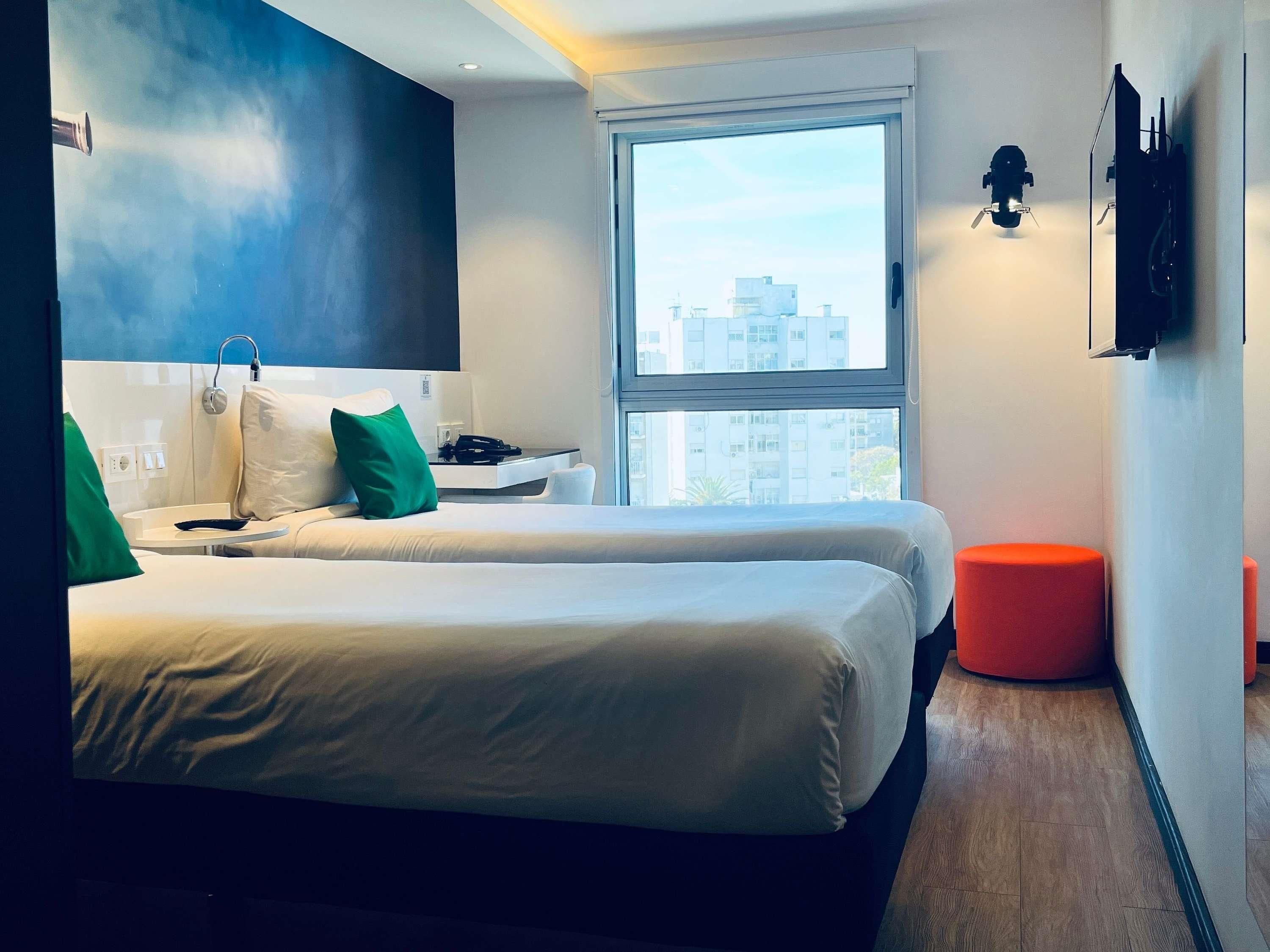 Ein modernes Hotelzimmer mit zwei Betten, einem großen Fenster und einem roten Pouf.