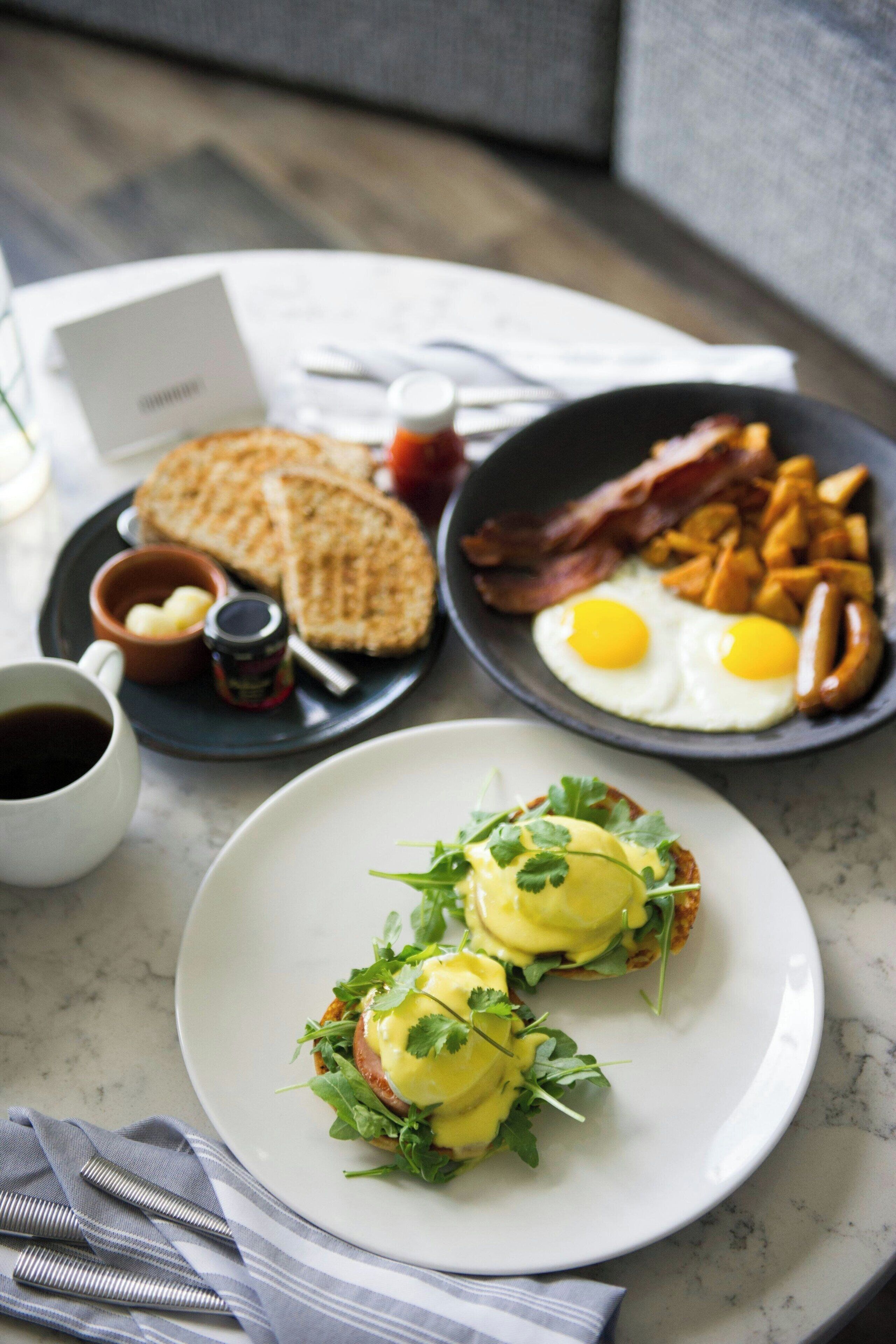 Un desayuno con tostadas, huevos, tocino y patatas ralladas en platos, una taza de café y un recipiente pequeño de mantequilla.