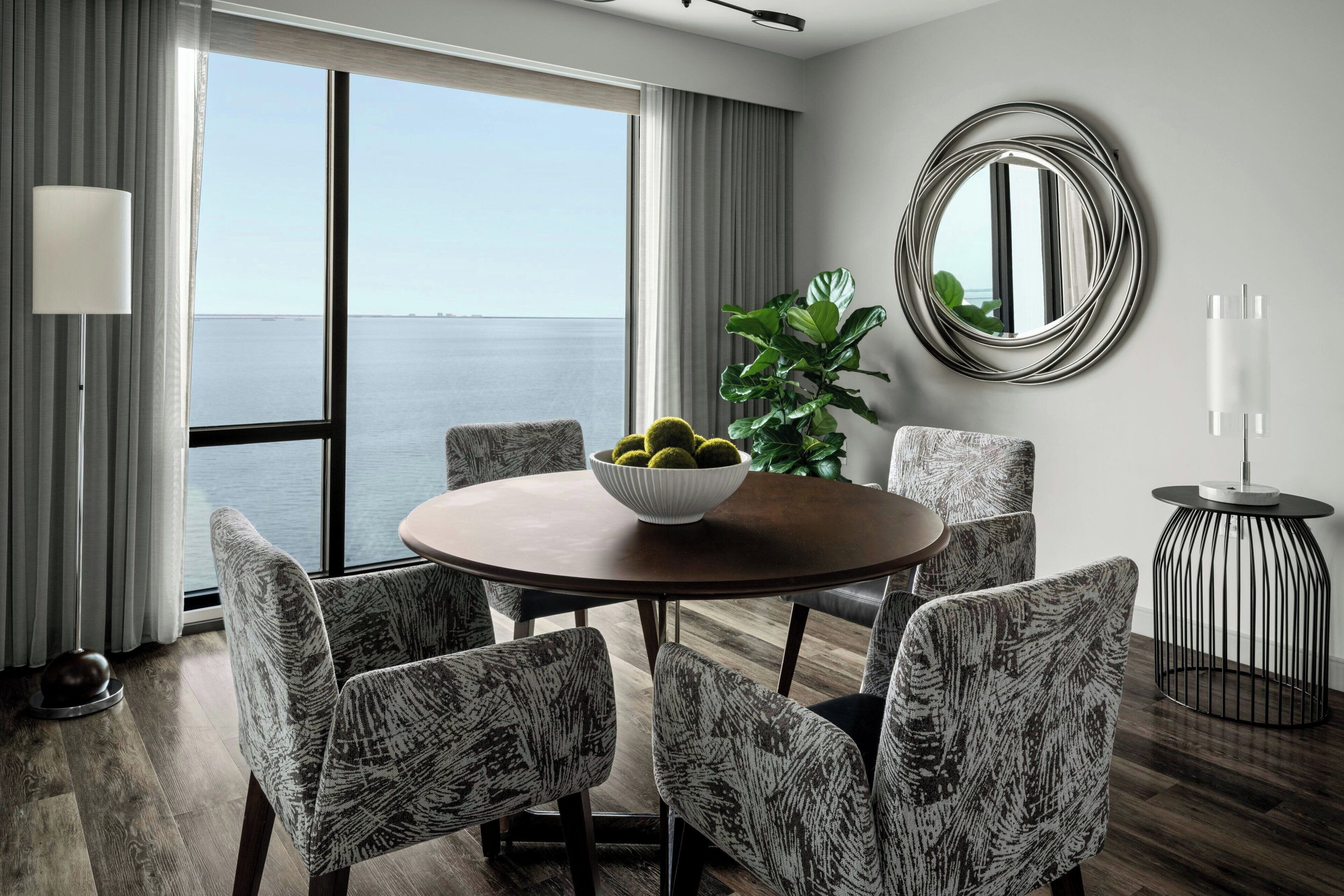 A dining area with a round table, six patterned chairs, a bowl of fruit, a potted plant, a round mirror, and a side table with candles.