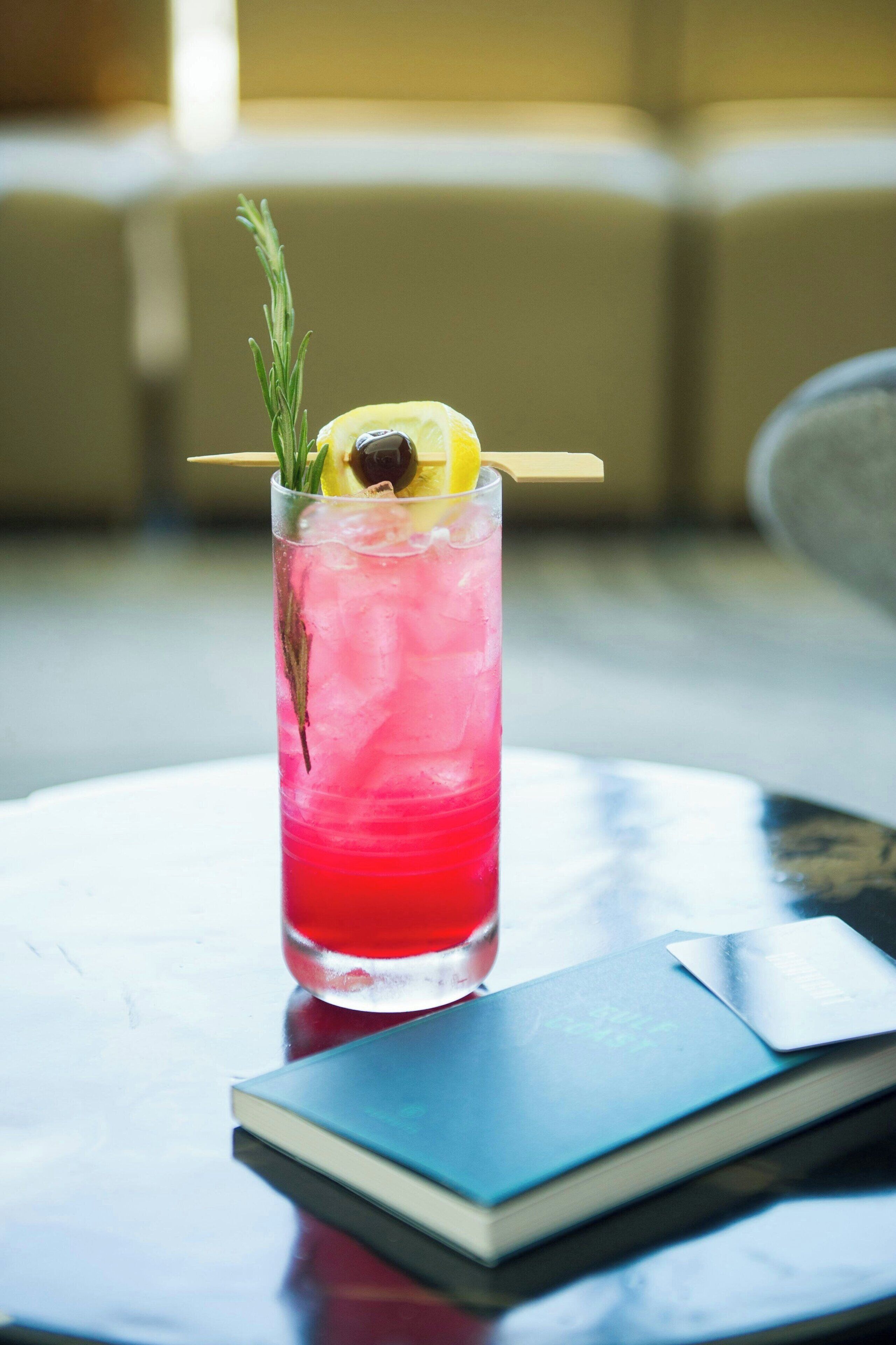Un verre à cocktail contenant une boisson rouge, agrémenté d’une tranche de citron, d’une olive et d’une branche de romarin, posé sur une table à côté d’un livre.