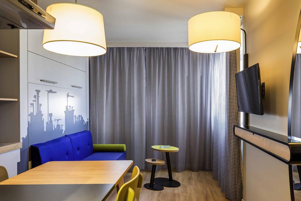 Une chambre d’hôtel moderne avec une œuvre d’art murale représentant un paysage urbain, un canapé bleu, une table à manger en bois, des chaises jaunes, une petite table ronde avec une lampe et un téléviseur fixé au mur.