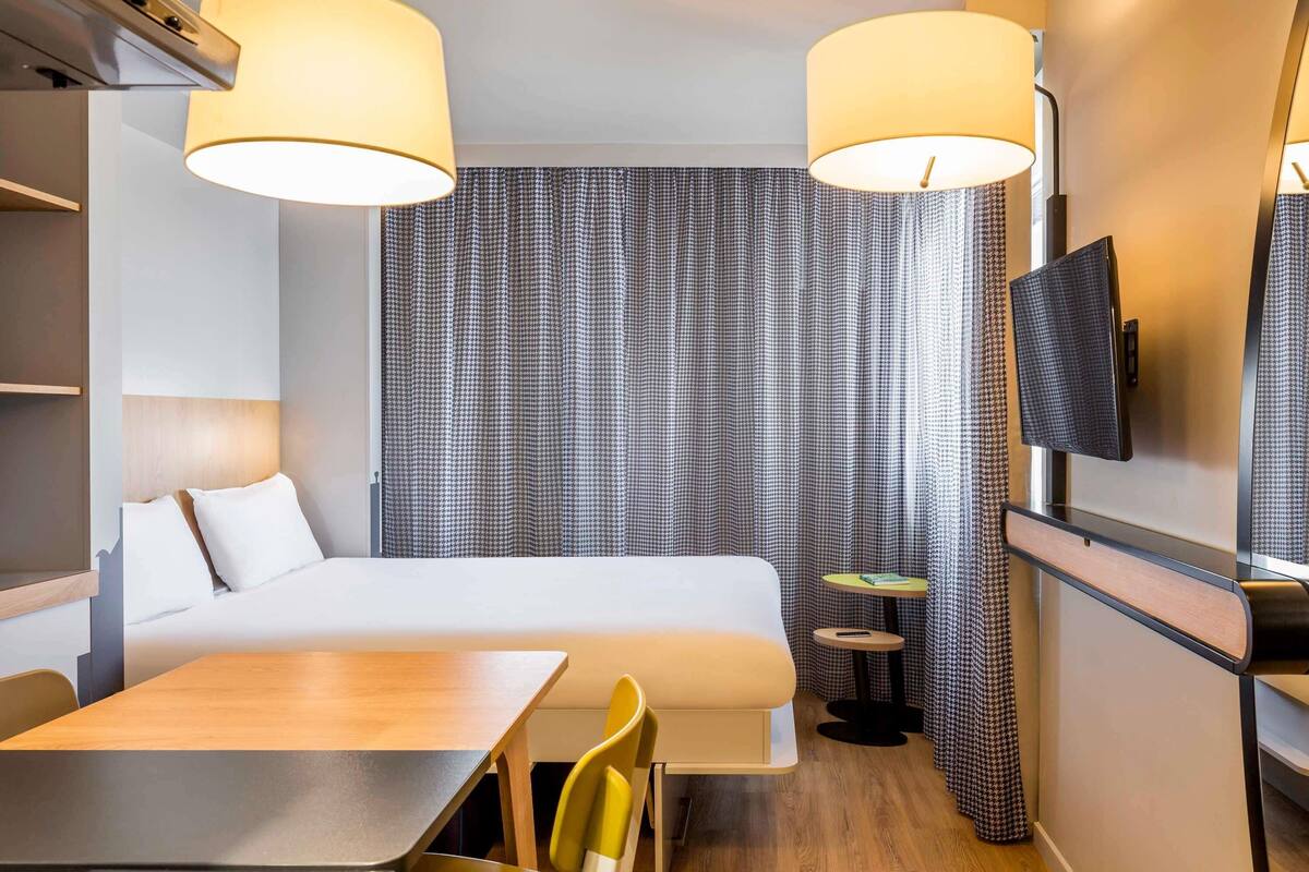 Quarto de hotel com uma cama, uma escrivaninha, duas cadeiras, uma mesinha, uma televisão e uma TV na parede.