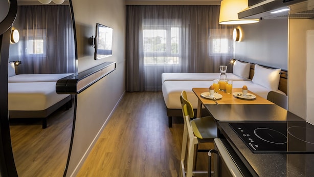 Kompakti hotellihuone, jossa on sänky, yöpöytä, pieni ruokailutila, sisäänrakennettu liesi ja suuri ikkuna, jossa on verhot.