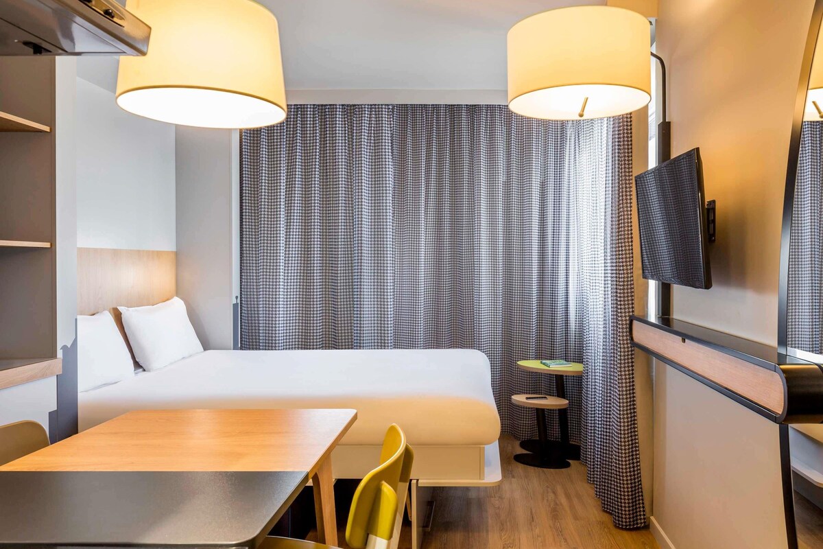 Habitación de hotel con cama, escritorio, dos sillas, una mesita, televisión y televisor de pared.