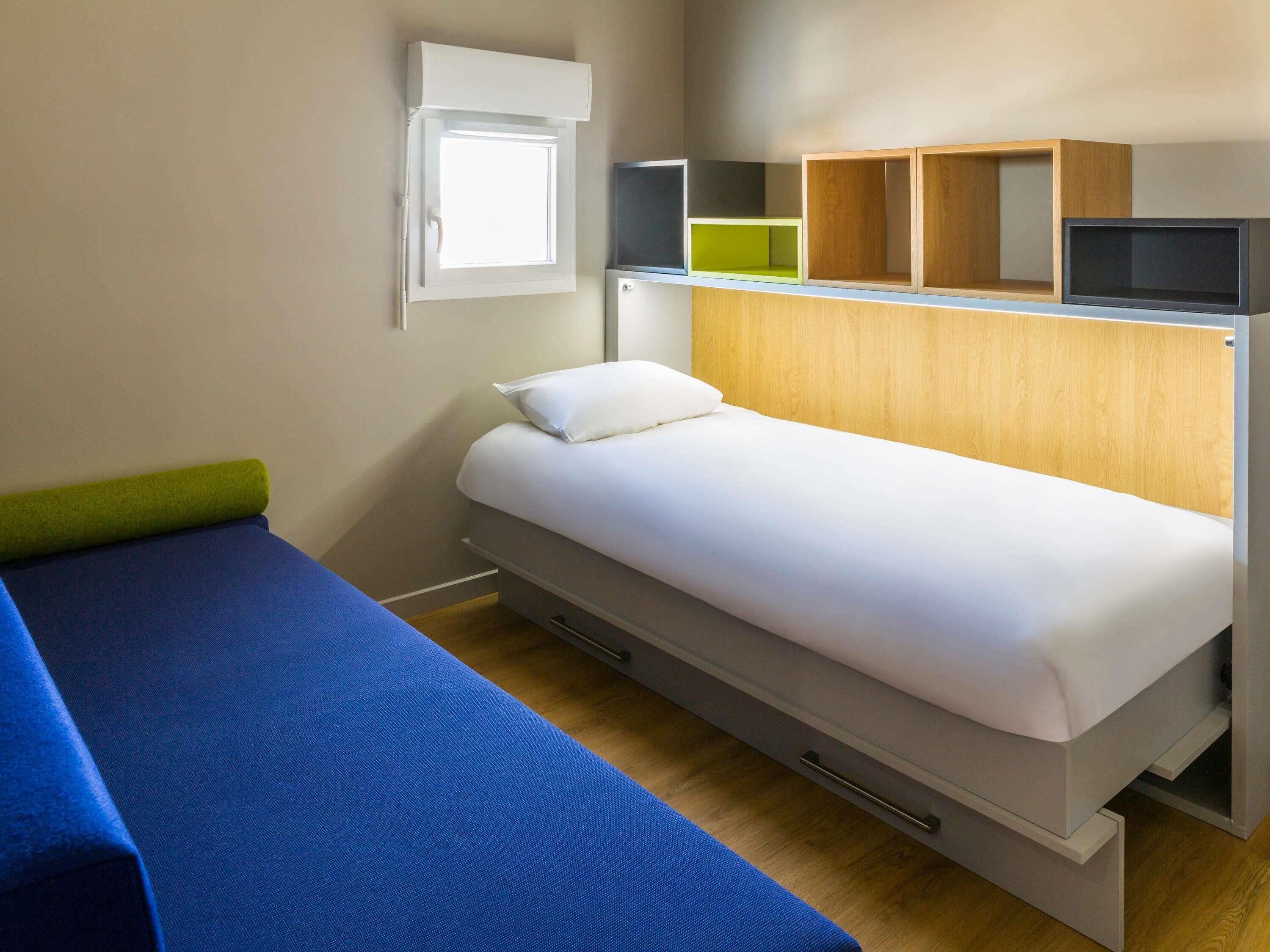 A single bed with a white mattress and a blue blanket, a small bedside table with a green object, a wooden headboard, and a window with a white blind.
