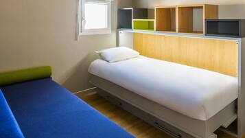 A single bed with a white mattress and a blue blanket, a small bedside table with a green object, a wooden headboard, and a window with a white blind.