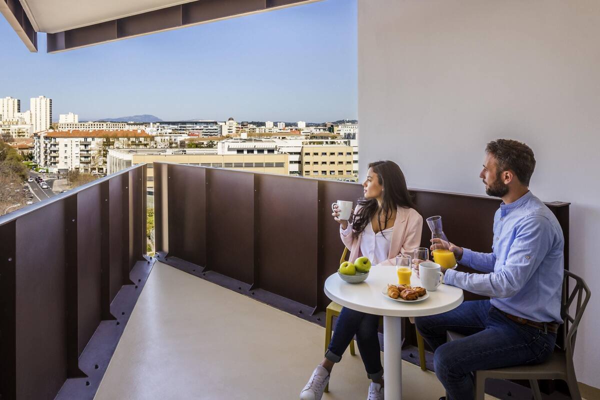 Uma varanda com vista para a cidade, uma mesa redonda com itens de café da manhã e uma pessoa segurando uma xícara.
