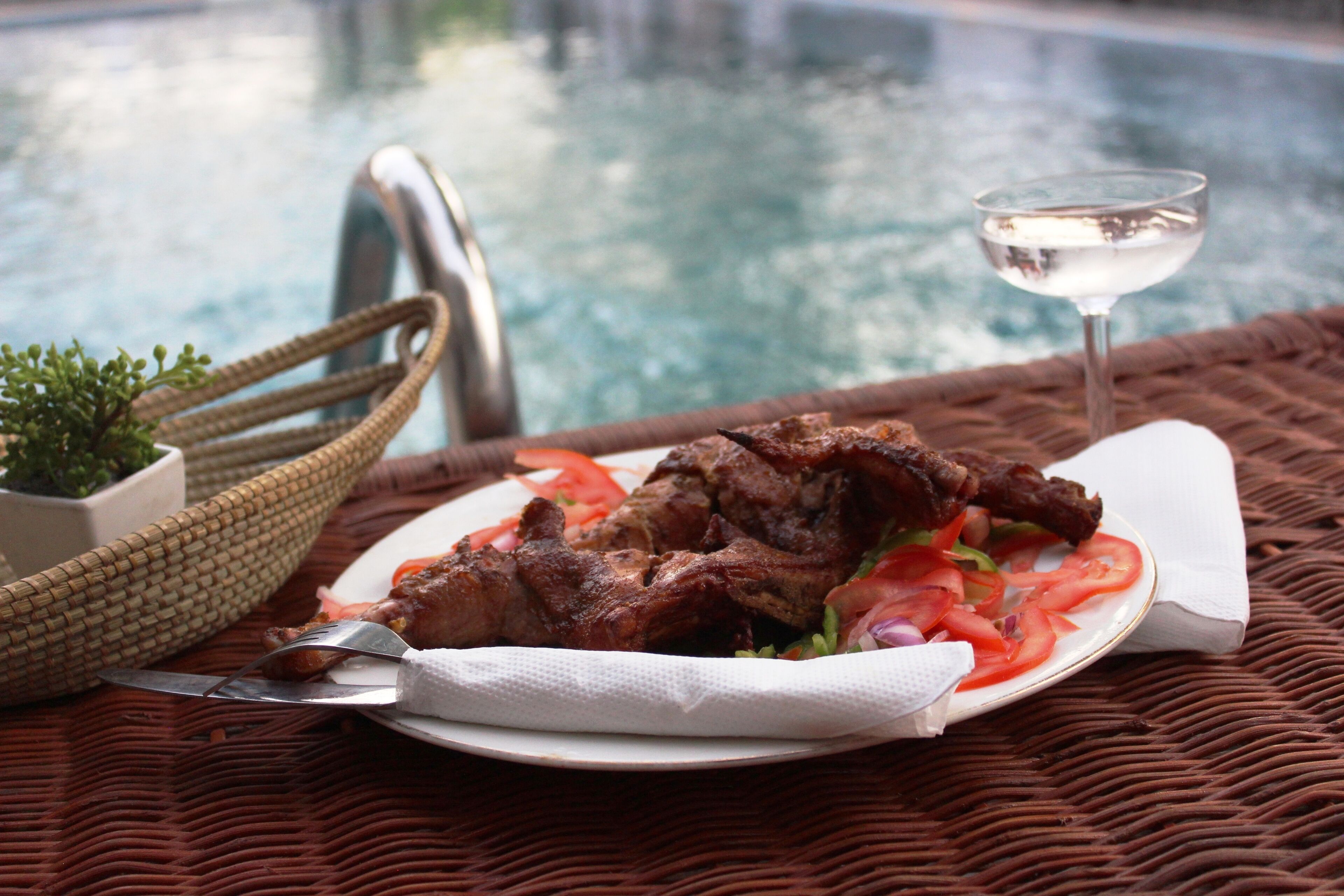 A plate of grilled meat with vegetables and a glass of water on a wicker table by a pool.