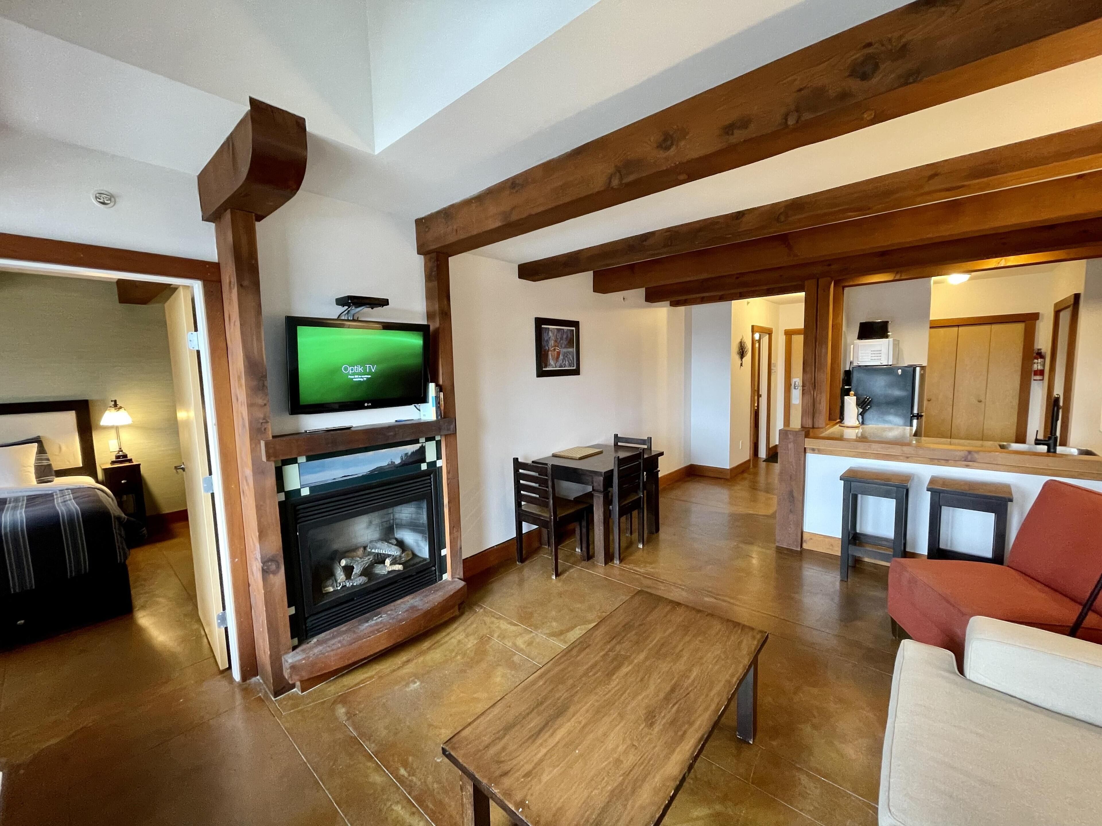 A living room with a fireplace, a television mounted above it, a dining area with a table and chairs, and a kitchen area with a counter and stools.