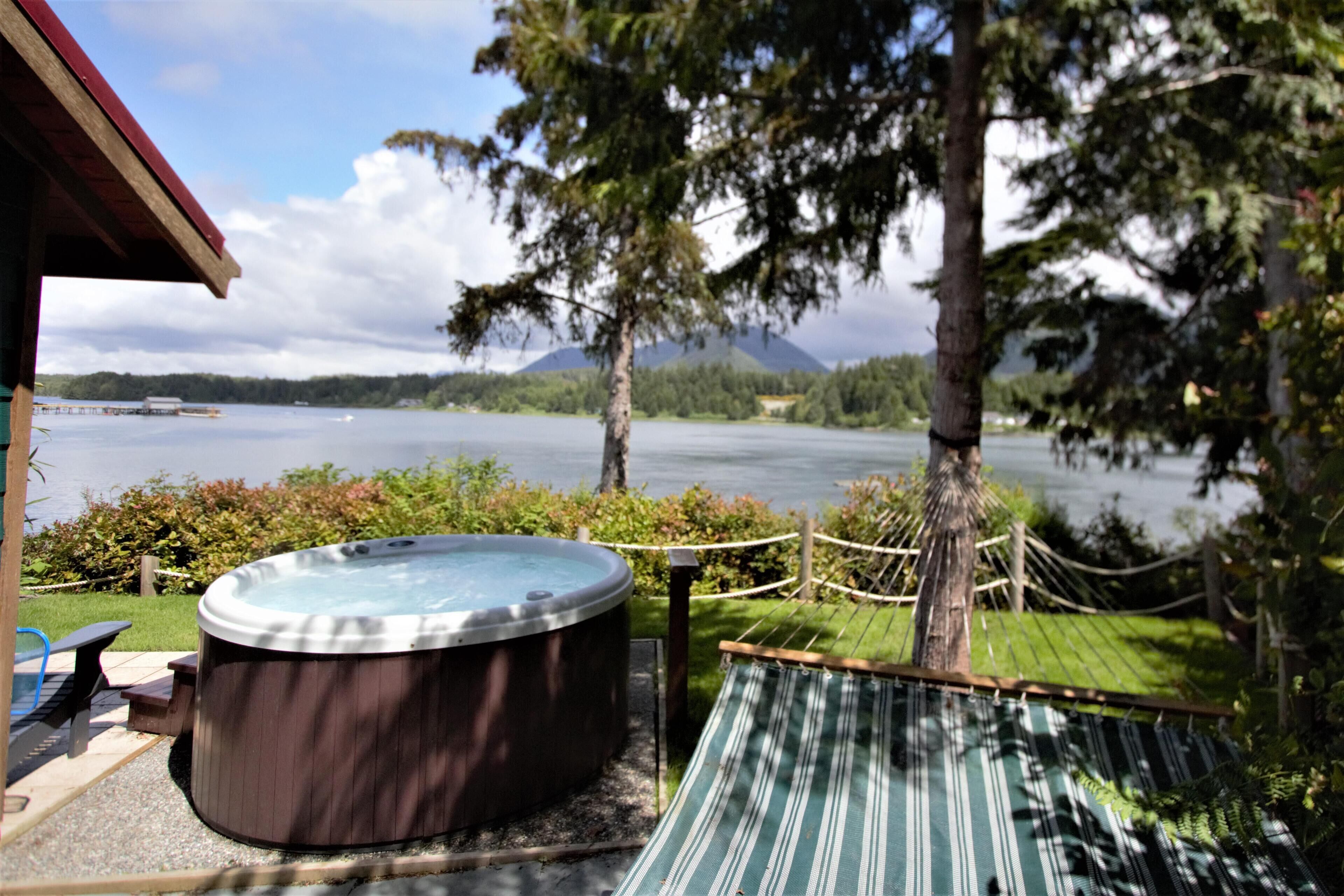 Un jacuzzi, una hamaca y una terraza de madera junto a un lago, con montañas al fondo.