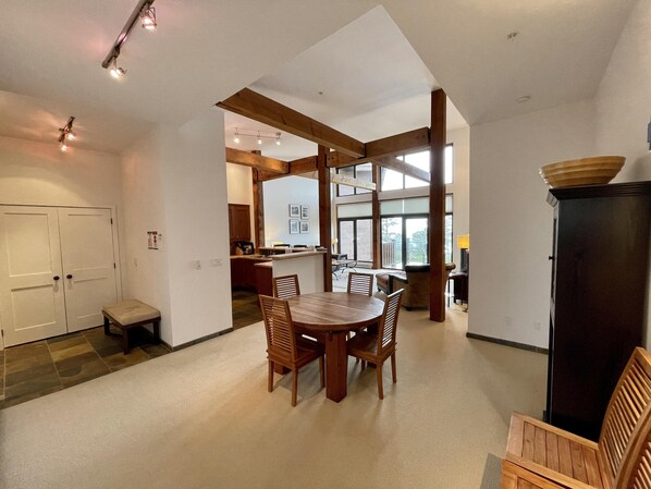 A spacious living area with a wooden dining table and chairs, a large window offering a view of the outdoors, and a kitchen area visible through an open doorway. - Big Beach Villa (Ucluelet)