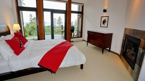 A bedroom with a large bed, a fireplace, a view of the outdoors, and a wooden dresser. - Pacific Rim Retreat (Ucluelet)